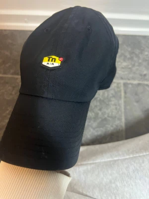 Svart Nike Tn Air keps - Svart keps från Nike med Tn Air-logga i gult och vitt framtill och gul swoosh bak. Justerbar snapback och tillverkad i Dri-FIT-material som andas. Snygg och sportig look, perfekt för dig som gillar streetwear och sneakers.