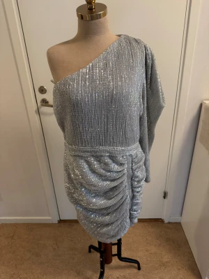 Glittrig silverklänning Lavish Alice - Superfin glittrig silverklänning från Lavish Alice med en ärm och snygg drapering över kjolen. One shoulder-modell som verkligen sticker ut och ger en cool vibe. Perfekt för dig som vill glänsa och synas! Stängs med dragkedja bak.