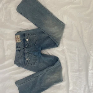 True Religion Becca Bootcut Jeans - Snygga ljusblå bootcut jeans från True Religion, modell Becca Mid Rise. Jeansen har klassiska fickor med lock och silverfärgade knappar, samt tydliga kontrastsömmar. Tillverkade i mjukt denim med en skön passform och cool faded effekt på benen. Nästan aldrig använda, hör av er om mått!❣️