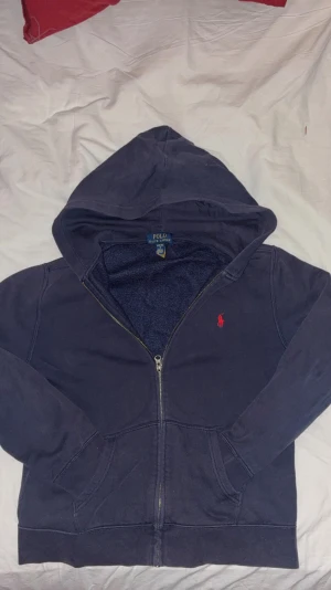 Mörkblå hoodie från Polo Ralph Lauren - Snygg mörkblå hoodie från Polo Ralph Lauren med röd broderad logga på bröstet. Tröjan har huva, dragkedja framtill och två fickor. Insidan är mjuk och fleecefodrad för extra komfort. Perfekt för chill dagar och en soft outfit.