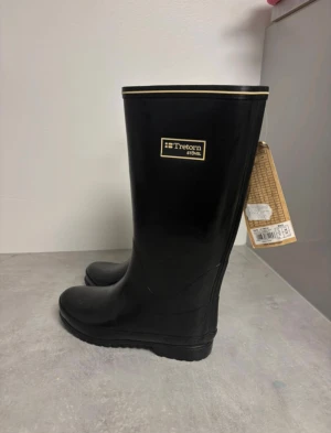 Svarta stövlar från Tretorn - Svarta höga stövlar från Tretorn med klassisk rund tå och platt sula. Tillverkade i slitstark syntet med diskret gulddetalj upptill. Perfekta för regniga dagar och har en stilren look som funkar till många outfits. Ny med prislapp ord pris 599kr