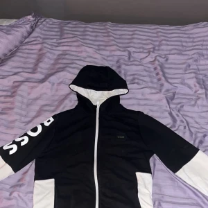 Svart och vit hoodie från BOSS - Snygg svart och vit hoodie från BOSS med dragkedja framtill och stor huva. Ärmarna har kontrasterande vita partier och BOSS-loggan tryckt i vitt på ena ärmen. Perfekt för dig som gillar streetwear och vill sticka ut lite extra.