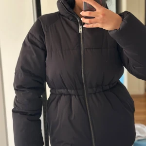 Svart pufferjacka med dragkedja - Säljer en svart pufferjacka med hög krage och hel dragkedja framtill. Jackan har markerad midja med resår och långa ärmar. Perfekt för kalla dagar när du vill hålla dig varm och ändå se snygg ut.