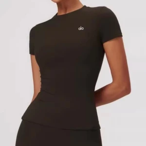 Svart topp från Alo Yoga - Säljer en svart, tight topp från Alo Yoga med korta ärmar och rund hals. Toppen har en stilren design med liten vit logga på bröstet och är tillverkad i ett stretchigt material som sitter snyggt mot kroppen.