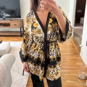 Svart och guld overshirt med leopardmönster - Säljer en sjukt snygg overshirt från Veromoda med leopardmönster och barockinspirerade detaljer i guld och svart. Jackan har omlottmodell med knyt i midjan, vida ärmar och v-ringad hals. Materialet är glansigt och känns lyxigt, perfekt för att sticka ut.
