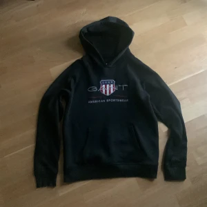Svart hoodie från GANT med tryck - Svart hoodie från GANT med stort tryck på bröstet och amerikansk sportswear-stil. Tröjan har huva, magficka och långa ärmar. Perfekt för dig som gillar klassisk streetwear och vill ha en snygg logga framtill.