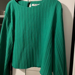 Grön plisserad blus från NA-KD - Säljer en snygg grön plisserad blus från NA-KD i storlek 38. Blusen har långa ärmar, rund halsringning och ett stilrent, strukturerat tyg med vertikala veck. Mörkare än tredje bilden. Helt ny oanvänd.
