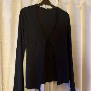 Svart långärmad bolerotopp BikBok - Svart långärmad bolerotopp från BikBok med djup v-ringning och knyt framtill. Toppen har vågiga kanter nertill och är tillverkad i ett mjukt, stretchigt material. Perfekt att styla över linne eller klänning för en trendig lager-på-lager-look.