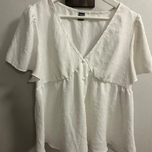 Vit blus med knappar från SHEIN - Supersöt vit blus från SHEIN med v-ringning och dekorativa knappar framtill. Blusen har korta ärmar, lätt struktur i tyget och en lös passform som ger en chill vibe. Perfekt till jeans eller kjol för en fräsch look.