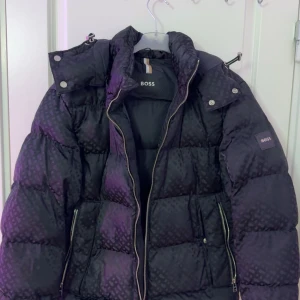 Riktigt fett pufferjacka från BOSS - Snygg svart pufferjacka från BOSS med diskret rutigt mönster. Jackan har hög krage, avtagbar huva med tryckknappar och dragkedjor på fickorna. Perfekt för kalla dagar och riktigt street i stilen.