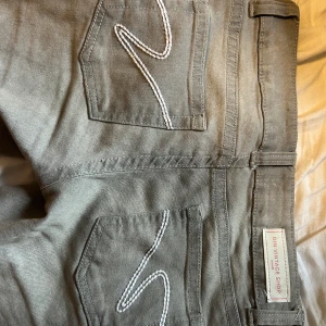 Grå bootcut jeans från Din Vintage Shop - Snygga grå jeans med bootcut passform från Din Vintage Shop. Byxorna har klassiska fem fickor, vita broderade detaljer på bakfickorna och dubbla knappar framtill. Perfekta för dig som gillar en stilren look med lite edge. Midja: 36cm, innerbenslängden: 81cm