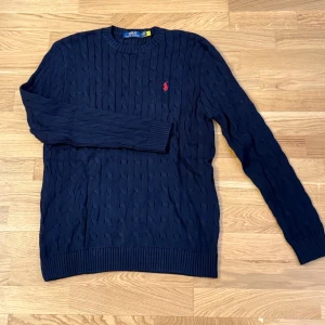 Mörkblå stickad tröja Polo Ralph Lauren - Kabelstickad mörkblå tröja från Polo Ralph Lauren med klassisk rund halsringning och röd broderad logga på bröstet. Tröjan är långärmad och har ribbade muddar vid ärmslut och nederkant. Perfekt för en stilren och tidlös look.