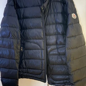 Marinblå Moncler dunjacka - Säljer en marinblå dunjacka från Moncler med klassisk quiltad design och dragkedja framtill. Jackan har lång ärm, diskret logotyp på ärmen och är tillverkad i glansig polyester. Perfekt för kyliga dagar och riktigt snygg till streetwear-stilen. Nypris ligger på 12 599