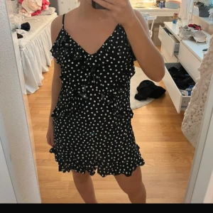 Svart prickig kort klänning med volang - Supersöt svart kort klänning med vita prickar och volangdetaljer. Klänningen har smala axelband, v-ringning och volanger längs både framstycke och fåll. 