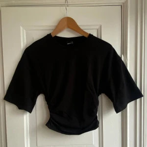 Svart croppad t-shirt Gina Tricot - Svart croppad t-shirt från Gina Tricot i mjuk bomull. Modellen har korta ärmar och en snygg rynkad detalj i midjan som ger en cool siluett. Perfekt för dig som gillar stilrena och trendiga plagg med lite edge.