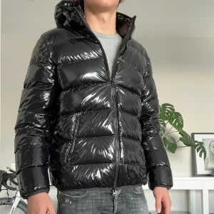 Svart pufferjacka från Grifoni - Säljer en svart, glansig pufferjacka från Grifoni med huva och dragkedja. Jackan har en riktigt fet look med breda, quiltade sektioner och två sidofickor. Perfekt för dig som vill sticka ut med en modern och urban stil.