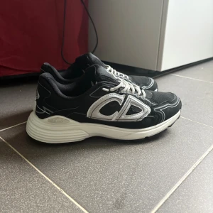 Svarta Dior B30 sneakers med vita detaljer - Säljer ett par svarta Dior B30 sneakers med vita och silvriga detaljer. Skorna har en chunky vit sula, mesh och skinnpaneler samt tydlig CD-logga på sidan. Klassisk snörning och sportig vibe, perfekta för dig som gillar lyxiga streetwear-vibbar. Helt nya använda 2 gånger bara 