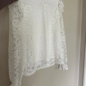 Vit spetsblus med långa ärmar - Superfin vit blus i spets med långärmad design och vackra blommiga detaljer i spetsmönstret. Blusen har rund halsringning och små volangdetaljer vid ärmslut och axlar. Perfekt för dig som gillar romantisk och feminin stil.