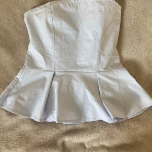 Vit peplum tubtopp från H&M - Säljer en vit peplum tubtopp från H&M i storlek S. Toppen är axelbandslös med figurnära passform upptill och utsvängd volang nertill. Perfekt för dig som vill ha en stilren och trendig look. Gjord i bomullsmix för en skön känsla.