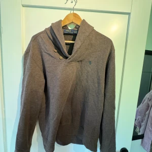 Brun tröja med knappar från Polo Ralph Lauren - Brun långärmad tröja från Polo Ralph Lauren med snygg krage och två knappar vid halsen. Tröjan har bruna mockadetaljer på armbågarna och en liten broderad logga på bröstet.