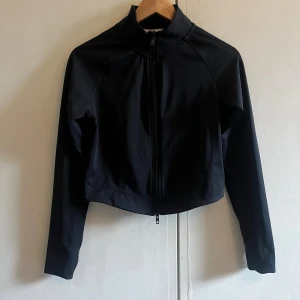 Svart croppad träningsjacka med dragkedja - Svart croppad träningsjacka med hög krage och hel dragkedja framtill. Jackan har långa ärmar med tumhål och är tillverkad i luftigt material som känns lätt att ha på sig. Aldrig använd!