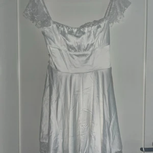 Vit klänning från Parthea - Supersöt vit kort klänning i satin-look från Parthea. Klänningen har spetsdetaljer vid axelbanden och över bysten, samt snörning i ryggen för extra snygg passform. 