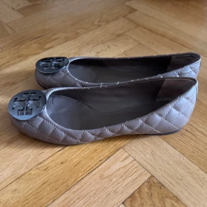 Ballerinaskor från Tory Burch storlek 39 - Snygga ballerinaskor från Tory Burch i beige quiltat skinn med rund tå. Skorna har en stor metallisk logga framtill. Storlek 39.