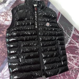 Svart dunväst från Moncler - Snygg svart dunväst från Moncler med glansig finish och klassisk logga på bröstet. Västen har hög krage, två dragkedjefickor och tydlig Moncler-detalj längs dragkedjan. Perfekt för lager-på-lager och streetstyle.