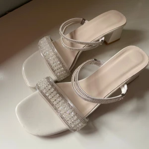 Vita sandaler med pärlor och klack - !Från Shein för många år sen! Vita sandaler med fyrkantig tå och klack. Remmarna är dekorerade med glittrande pärlor och strassdetaljer som ger extra bling. Kan inte garantera någon kvalitet, använda endast ett fåtal gånger.