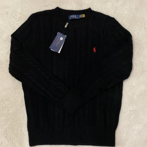 Svart kabelstickad tröja Polo Ralph Lauren - Svart kabelstickad tröja från Polo Ralph Lauren med klassisk röd broderad logga på bröstet. Tröjan har rund halsringning, ribbade muddar och är långärmad. Perfekt för dig som gillar stilren och tidlös design.