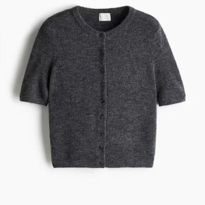 Mörkgrå stickad kofta med kort ärm - Mysig mörkgrå stickad kofta med korta ärmar och rund hals. Koftan har knappar hela vägen framtill och ribbade kanter vid ärmslut och nederkant. Perfekt att slänga över en t-shirt eller linne för en chill look.