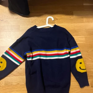 Mörkblå stickad tröja med smileys - Mörkblå stickad tröja med färgglada ränder över bröstet och armarna i blått, gult, rosa, rött, grönt och vitt. På båda ärmarna finns stora gula smiley-ansikten. Tröjan har rund hals och långa ärmar. Perfekt för dig som gillar färg och lekfull stil.