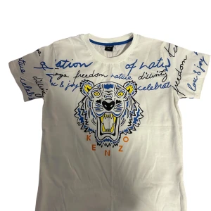 Vit Kenzo t-shirt med tigertryck - Snygg vit t-shirt från Kenzo med ikoniskt tigertryck framtill och blå samt svarta handskrivna texter över hela framsidan och ärmarna. Klassisk passform och rund halsringning. Perfekt för dig som vill sticka ut med en cool streetstyle-look. Till pojkar 11-12 år 