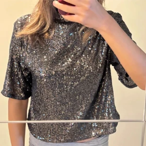 Svart glittrig topp med paljetter - Säljer en svart glittrig topp täckt av paljetter. Toppen har korta ärmar och en lite lös passform, perfekt för att sticka ut. Den höga halsen ger en cool vibe och materialet känns festligt och trendigt.