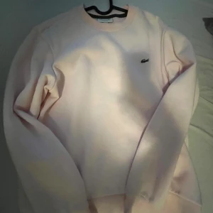 Ljusrosa sweatshirt från Lacoste - Säljer en ljusrosa sweatshirt från Lacoste med klassisk rund hals och det ikoniska krokodilloggan broderad på bröstet. Tröjan har långärmade och är gjord i mjuk bomull som är skön att ha på sig. Perfekt för chill dagar eller när du vill ha en clean look. Helst mötas upp