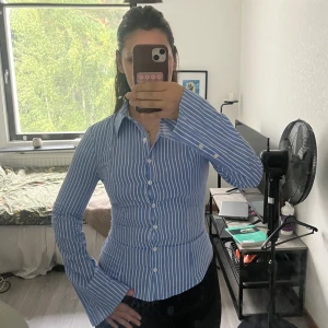 Blåvit randig skjorta från Bershka - Snygg blå skjorta med vita ränder från Bershka. Skjortan har klassisk krage, långa ärmar med breda manschetter och knäppning framtill. Figurnära passform som ger en clean och fräsch look. Perfekt för dig som gillar stilrena vibbar. Aldrig använd!