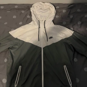 Nike vindjacka med huva, vit och grå - Nike vindjacka i vitt och mörkgrått med dragkedja framtill och två fickor med dragkedja. Jackan har huva med snörning och meshfoder på insidan. Perfekt för blåsiga dagar och snygg streetstil.