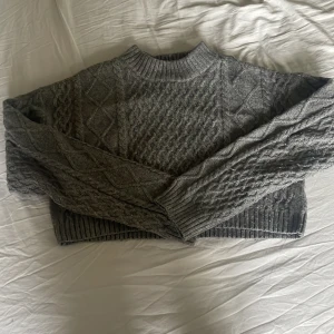 Grå stickad tröja från H&M - Mysig grå stickad tröja från H&M i storlek XS. Tröjan har ett klassiskt kabelstickat mönster, ribbade muddar och hög hals. Perfekt för kyliga dagar och enkel att matcha med jeans eller kjol.