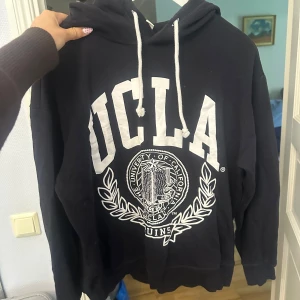 Mörkblå UCLA hoodie från H&M XS - Mörkblå hoodie från H&M i storlek XS med stor vit UCLA-print på framsidan och snörning i huvan. Tröjan har långa ärmar och ribbade muddar. Materialet är mjuk bomull, perfekt för chill dagar. Klassisk college-vibe med snygg logga framtill.