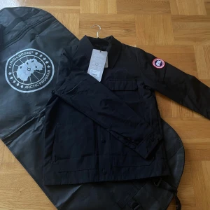 Canada Goose Burnaby - Svart Burnaby från Canada Goose med klassisk krage, stora fickor framtill och ikoniskt märke på ärmen. Jackan är tillverkad i polyester och har en stilren look som passar perfekt till lager-på-lager. Snygg och enkel att matcha med det mesta. En väldigt unik jacka för dig som vill ha något unikt! Storlek XS men passar som S/M
