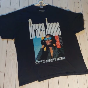 Grace Jones svart t-shirt 2XL - Svart t-shirt med stor Grace Jones-tryck framtill, texten 'Slave to Nobody's Rhythm' och färgglada detaljer. Oversized passform och klassisk rund hals. Tillverkad i mjuk bomull, perfekt för dig som gillar statement-plagg och ikoniska motiv.