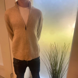 Beige stickad kofta med dragkedja från Ralph lauren - Beige stickad kofta med hel dragkedja och hög krage. Koftan har ribbade muddar vid ärmslut och nederkant, vilket ger en snygg och avslappnad look. Perfekt för lager-på-lager under kyliga dagar.