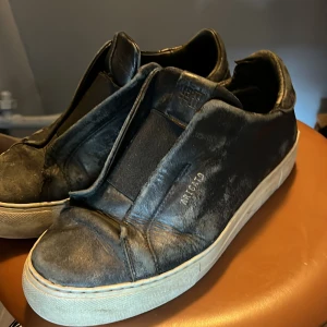 Arigato - Svarta sneakers från Axel arigato  i skinn med vit platt sula och minimalistisk design. Skorna har en stilren look utan synlig snörning och den ikoniska guldpräglade nummerkoden på sidan. Perfekta för dig som gillar clean och modern stil.