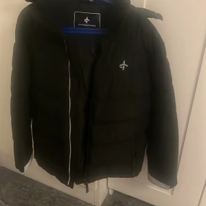 Svart pufferjacka från Cross Sportswear - Svart pufferjacka från Cross Sportswear med huva och diskret logga på bröstet och huvan. Jackan har dragkedja framtill och är vadderad för extra värme. Perfekt för kalla dagar och har en stilren look som funkar till det mesta.