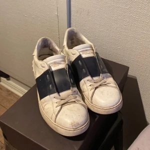 Valentino Garavani Open sneakers - Snygga Valentino Garavani Open sneakers i vitt skinn med bred svart rem över mitten. Klassisk rund tå, platt sula och snörning framtill. Perfekta för dig som gillar exklusiva och trendiga sneakers med en unik touch.