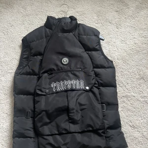 Svart dunväst från Trapstar London - Säljer en svart dunväst från Trapstar London med stor logga broderad både fram och bak. Västen har en unik asymmetrisk dragkedja, stor ficka framtill med knapp och dragkedja samt hög krage. Tillverkad i 100% polyester och har en riktigt streetig vibe. Passar både S och M