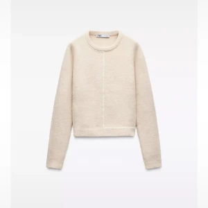 Beige stickad tröja från Zara - Beige stickad tröja från Zara med rund halsringning och lång ärm. Tröjan har en unik vit linje som går vertikalt och horisontellt framtill och baktill, vilket ger en stilren och modern look. Perfekt till jeans eller kjol för en clean vibe. Fint skick, något nopprig står XL men köpt utomlands så den motsvarade M, passar mer S
