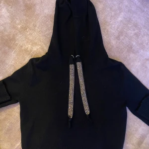 Svart hoodie med glittriga snören från Zara - Svart stickad hoodie från Zara med långa ärmar och huva. Tröjan har breda, glittriga snören framtill som ger en extra cool touch. Perfekt för dig som vill ha något bekvämt men ändå sticker ut lite. Mjuk och skön kvalitet.