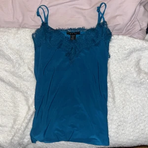 Blå silkes linnetopp med spets från Amisu - Säljer en blå silkes linnetopp från Amisu i storlek XS. Toppen har tunna axelband och v-ringning med dekorativ spets längs kanten. .
