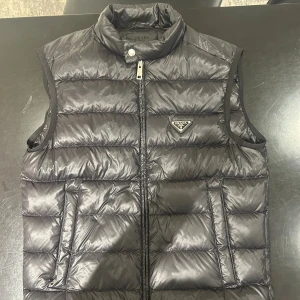 Svart dunväst från Prada - Snygg svart dunväst från Prada med quiltad design och hög krage. Västen har dragkedja framtill, två fickor och den ikoniska Prada-loggan på bröstet. Perfekt för lager-på-lager och ger en clean, lyxig look.